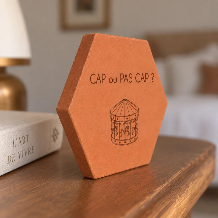 Tomette gravée « Cap ou pas cap ? » avec carrousel, vue de côté sur une table en bois près d’un livre.