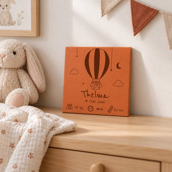 Carreau de naissance personnalisé Thelma, montgolfière et prénom gravés, sur une commode, près d’un lapin en peluche et de fanions.