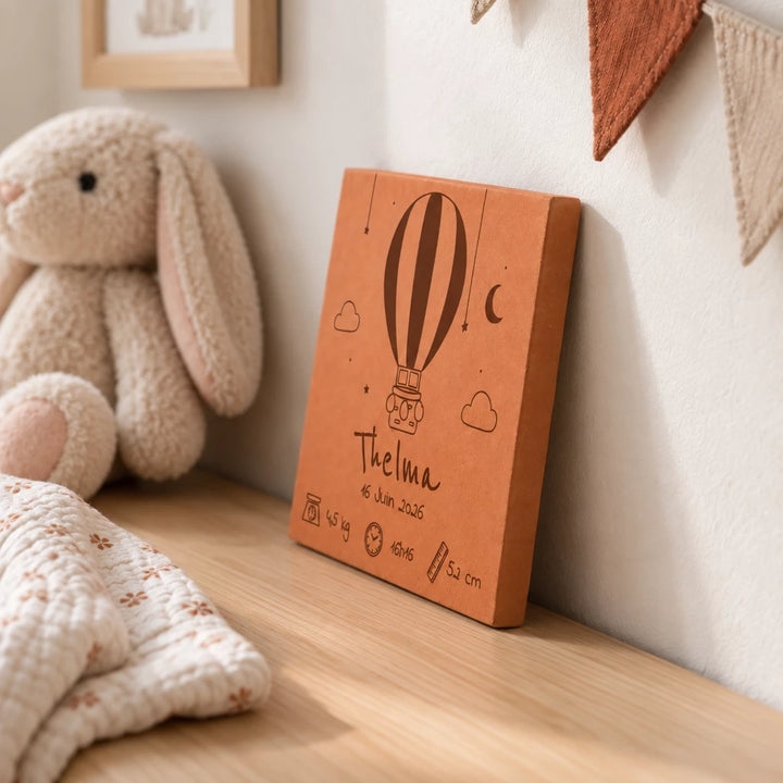 Carreau de naissance personnalisé Thelma, montgolfière et prénom gravés, vu de côté sur une commode près d’un lapin en peluche.