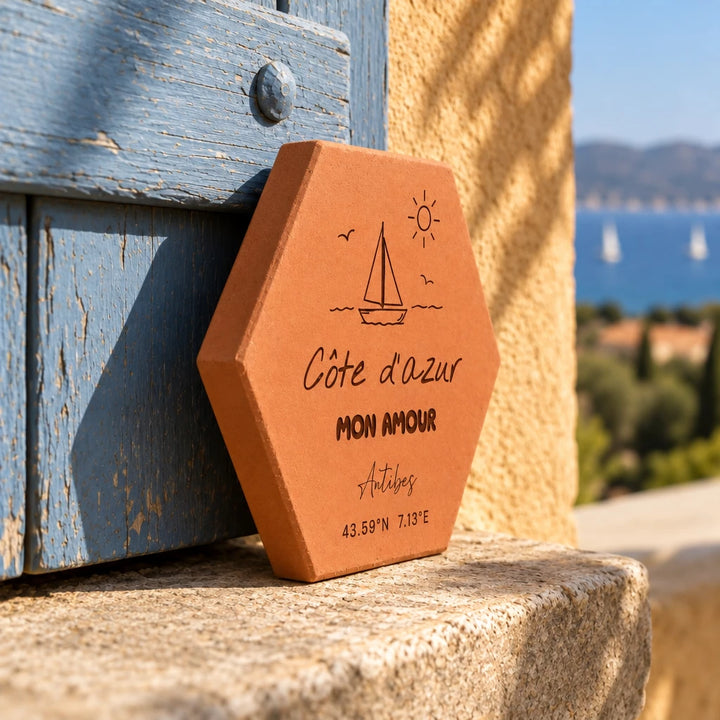 Tomette souvenir personnalisée « Côte d’Azur mon amour », Antibes, voilier et coordonnées gravés, vue de côté sur un rebord en pierre contre un volet bleu.
