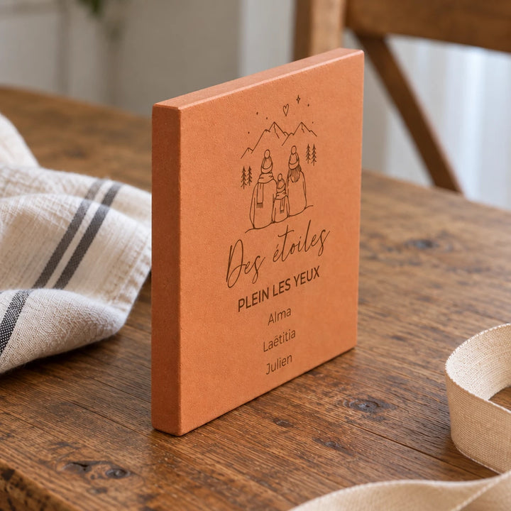 Carreau personnalisé « Des étoiles plein les yeux », famille en montagne et trois prénoms gravés, vu de côté sur une table en bois, tranche visible près d’un ruban.