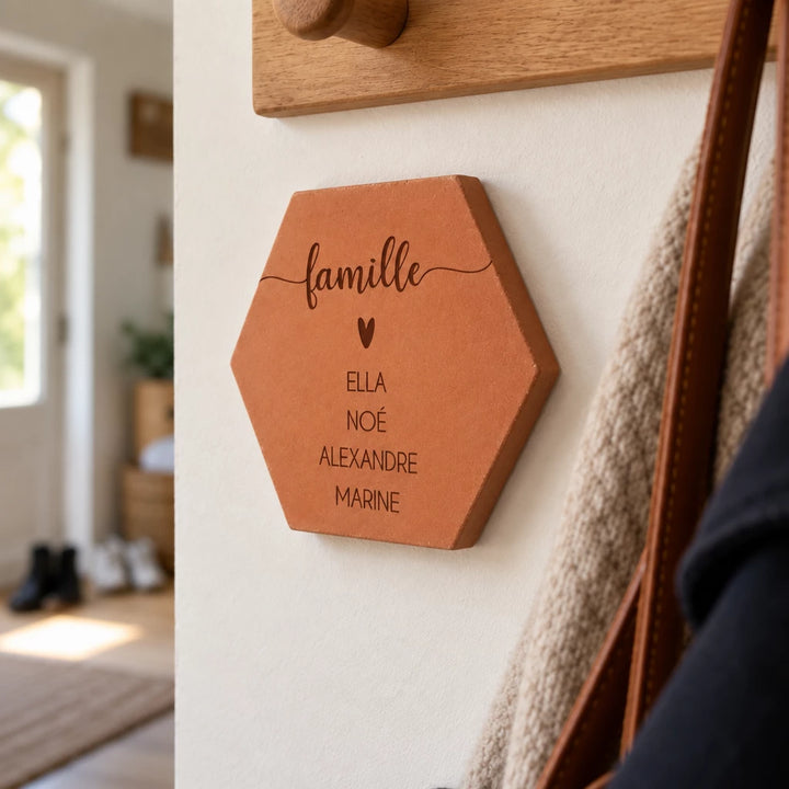 Tomette personnalisée « famille », cœur et quatre prénoms gravés, vue de biais sous un porte-manteau en bois.