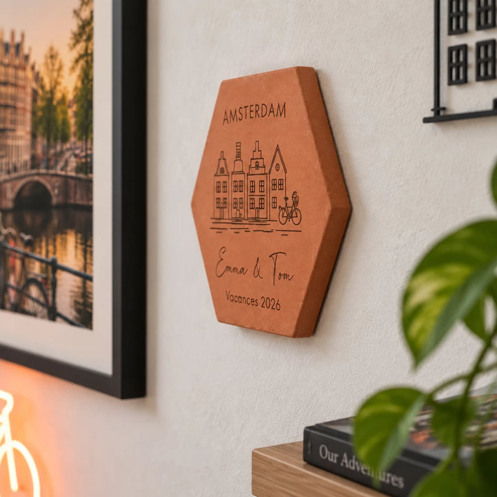 Tomette souvenir personnalisée Amsterdam, maisons, vélo, Emma et Tom et année gravés, vue de côté au mur près d’une plante et d’un album de voyage.