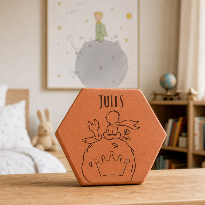 Tomette personnalisée Jules, petit prince et renard sur une planète gravés, sur une table en bois devant une affiche et un lit.