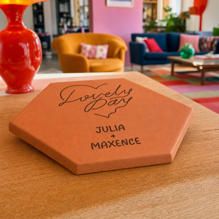 Tomette de couple personnalisée « Lovely Day », cœur et prénoms Julia et Maxence gravés, posée à plat sur une table en bois près d’une lampe rouge.