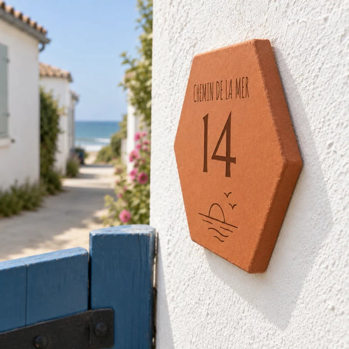 Plaque de maison personnalisée 14 chemin de la Mer, soleil et vagues gravés, vue de côté près du portillon bleu dans une ruelle côtière.