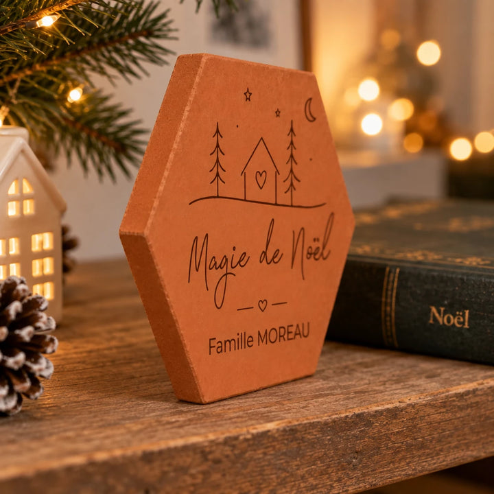 Tomette de Noël personnalisée « Magie de Noël », Famille Moreau et petite maison gravées, vue de côté près de branches de sapin et d’un livre de Noël.
