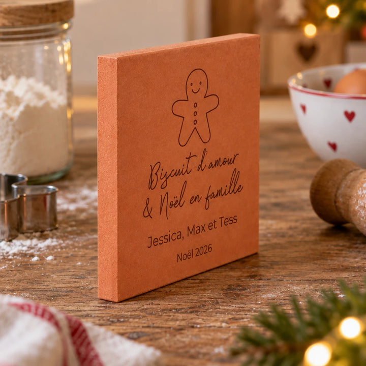 Carreau de Noël personnalisé « Biscuit d’amour », bonhomme pain d’épices et trois prénoms gravés, vu de côté sur un plan de travail décoré de branches de sapin.