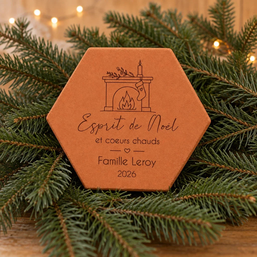 Tomette Esprit de Noël avec un nom de famille, posée parmi des branches de sapin et une guirlande lumineuse.