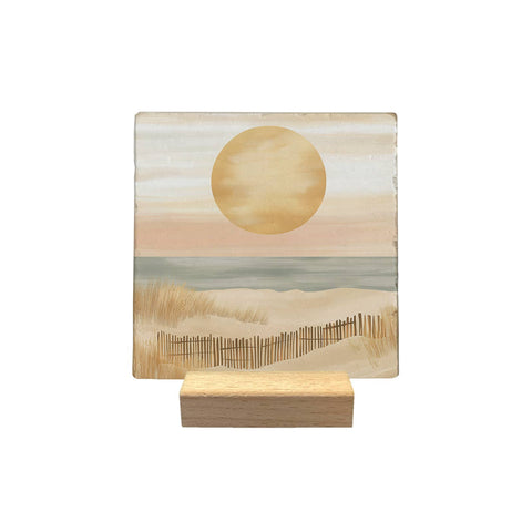 Dunes decorative tile, with a golden sun, seashore and a fence in the sand, displayed on a small wooden stand against a white background.