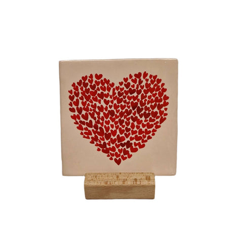 Decorative tile with a large heart made up of small red hearts, displayed alone on a small wooden stand against a white background.