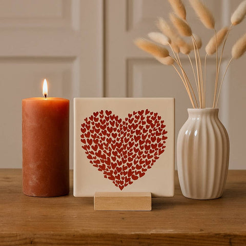 Decorative tile with a large heart made up of small red hearts, on a wooden stand beside a candle and a vase of dried flowers.