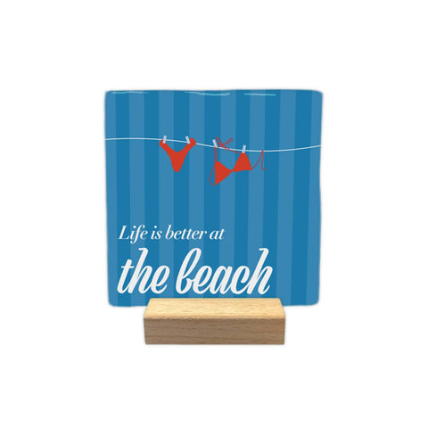 “Life is better at the beach” decorative tile, with red swimsuits and blue stripes, displayed on a small wooden stand against a white background.