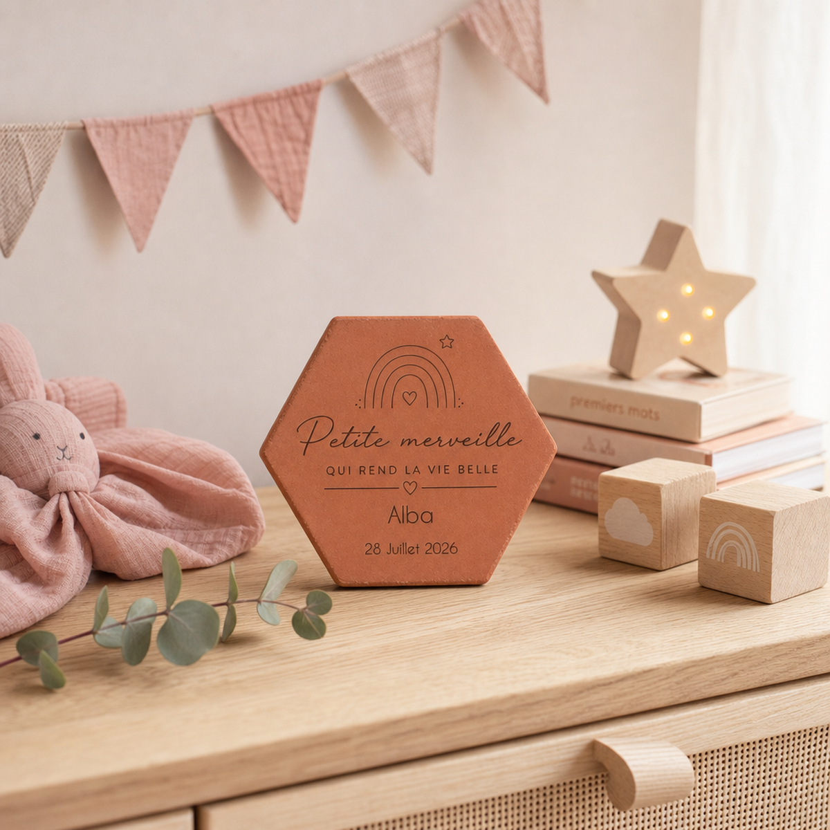 Tomette de naissance personnalisée Alba, arc-en-ciel et « Petite merveille » gravés, sur une commode près d’une étoile lumineuse, de livres et de cubes.