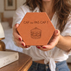 Tomette gravée « Cap ou pas cap ? » avec carrousel, présentée entre deux mains devant une chemise blanche.