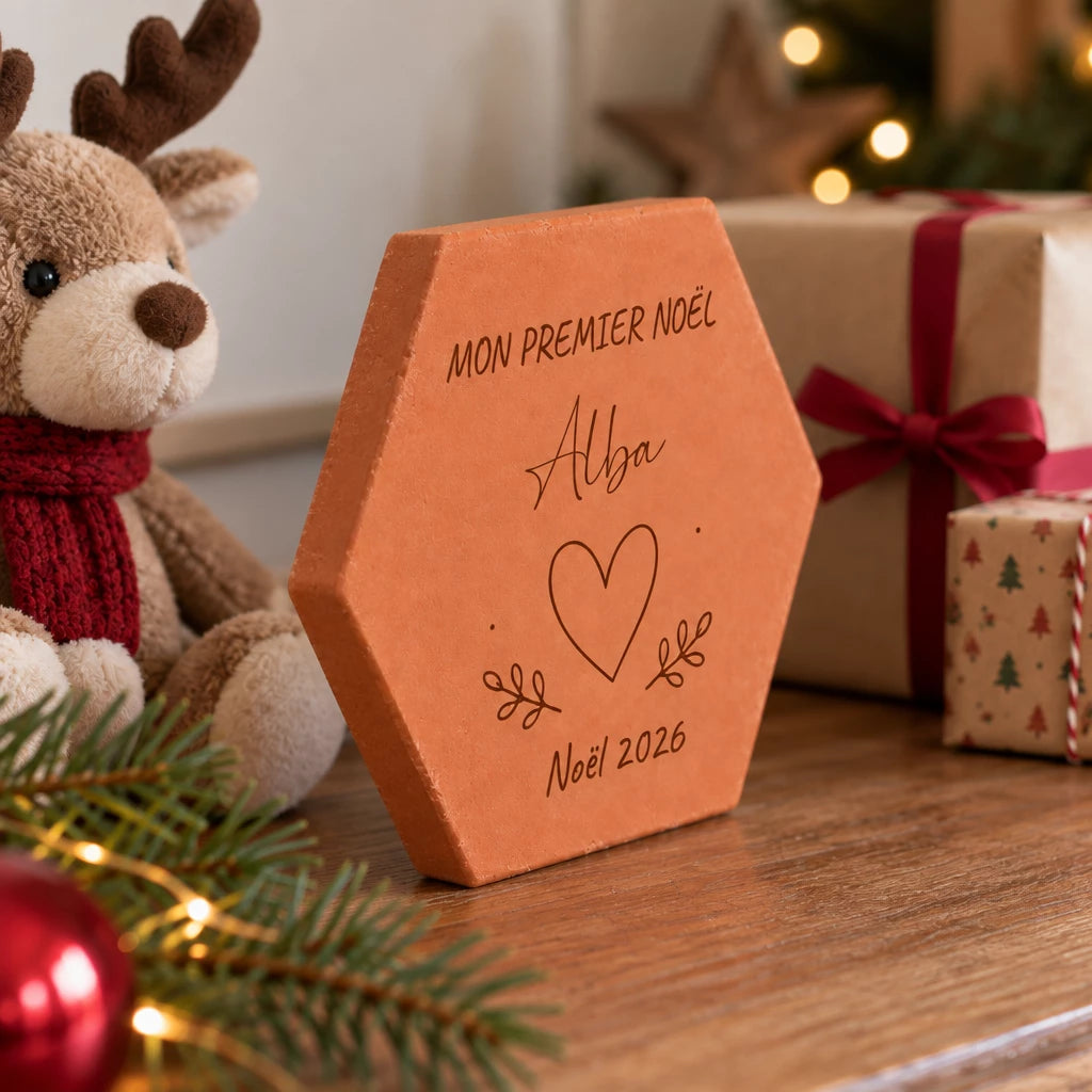 Tomette personnalisée « Mon premier Noël », Alba, cœur et année gravés, vue de côté près du renne, des cadeaux et d’une branche de sapin.