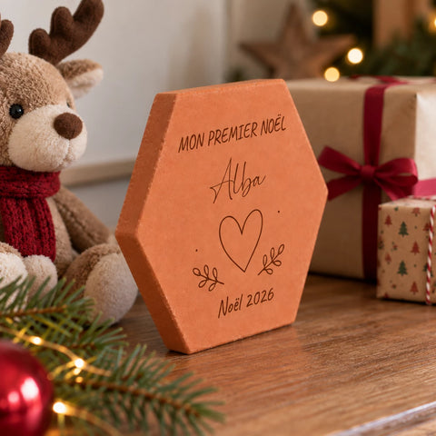 Tomette personnalisée « Mon premier Noël », Alba, cœur et année gravés, vue de côté près du renne, des cadeaux et d’une branche de sapin.