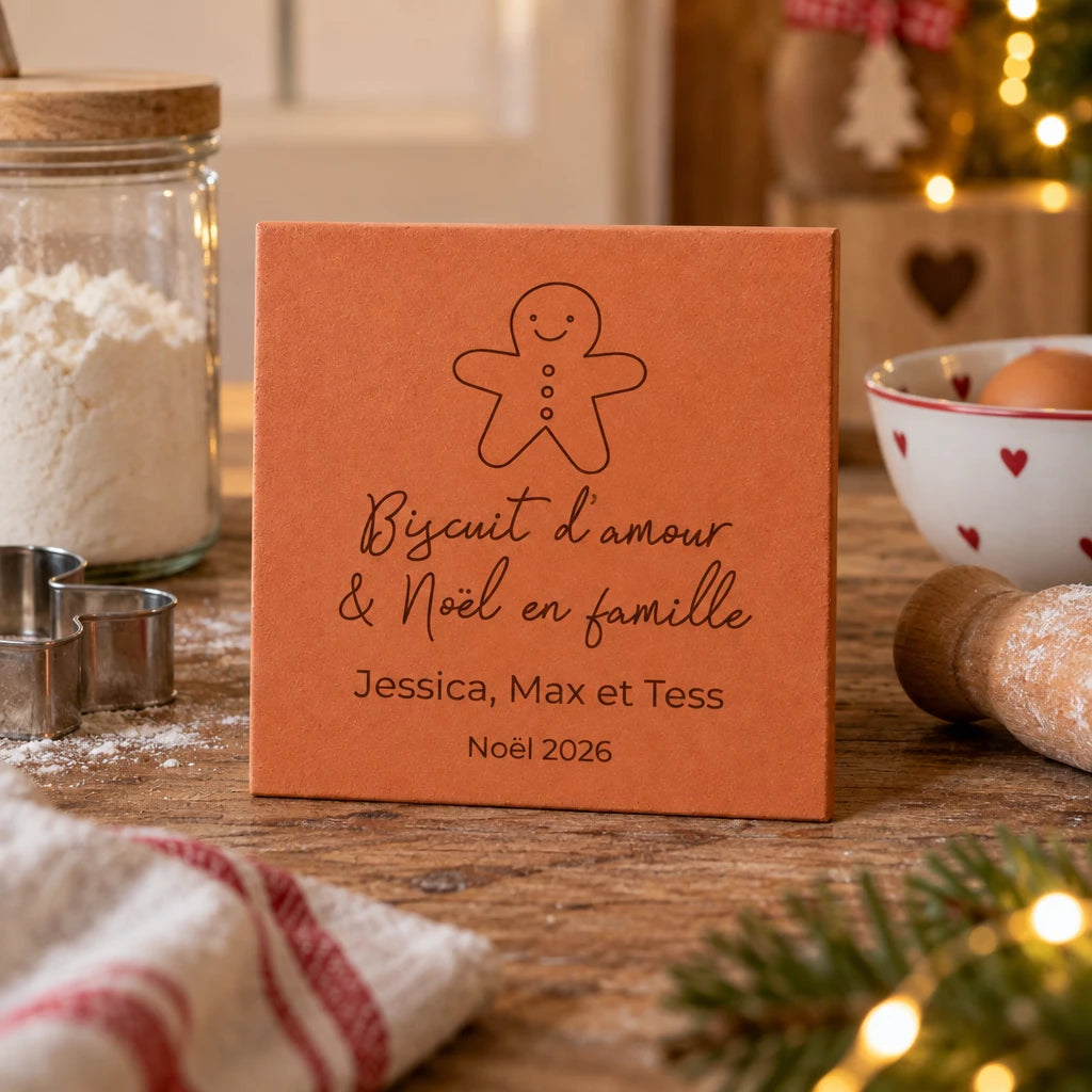 Carreau de Noël personnalisé « Biscuit d’amour », bonhomme pain d’épices et trois prénoms gravés, près d’un bocal, d’emporte-pièces et d’un rouleau à pâtisserie.