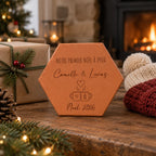 Tomette personnalisée « Notre premier Noël à deux », Camille et Lucas et deux tasses gravés, sur une table près d’un cadeau et de bonnets, devant une cheminée.
