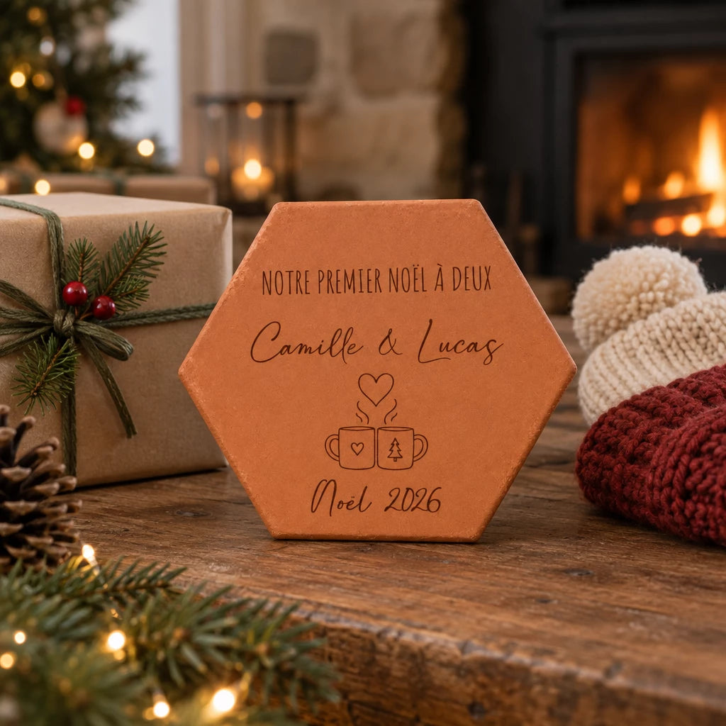 Tomette personnalisée « Notre premier Noël à deux », Camille et Lucas et deux tasses gravés, sur une table près d’un cadeau et de bonnets, devant une cheminée.