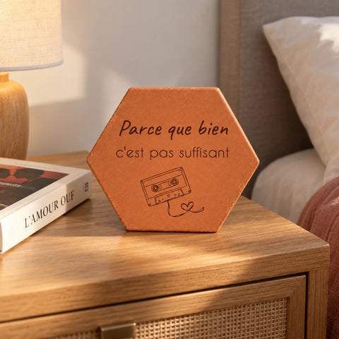 Terracotta tile engraved with “Parce que bien c’est pas suffisant”, an audio cassette and heart-shaped tape, on a bedside table beside a lamp and a book.