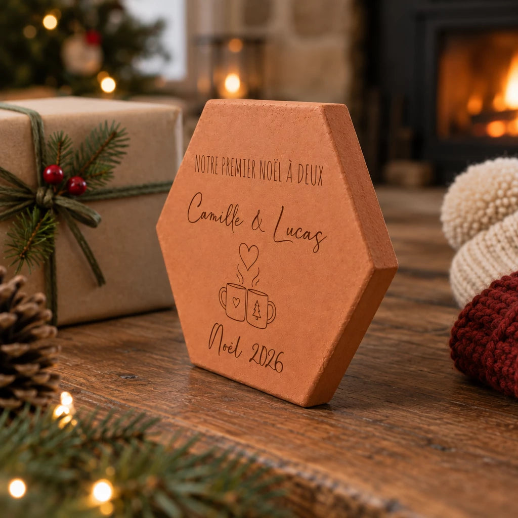 Tomette personnalisée « Notre premier Noël à deux », Camille et Lucas et deux tasses gravés, vue de côté près d’un paquet cadeau et de branches de sapin.