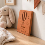 Carreau de naissance personnalisé Thelma, montgolfière et prénom gravés, vu de côté sur une commode près d’un lapin en peluche.