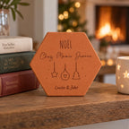 Tomette personnalisée « Noël chez Mamie Jeanne », Louise et Jules et décorations gravés, sur une table près de livres, d’une tasse et d’une cheminée allumée.