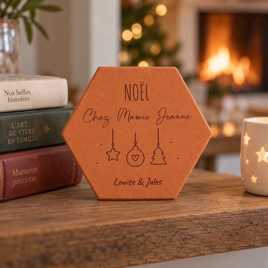 Tomette personnalisée « Noël chez Mamie Jeanne », Louise et Jules et décorations gravés, sur une table près de livres, d’une tasse et d’une cheminée allumée.