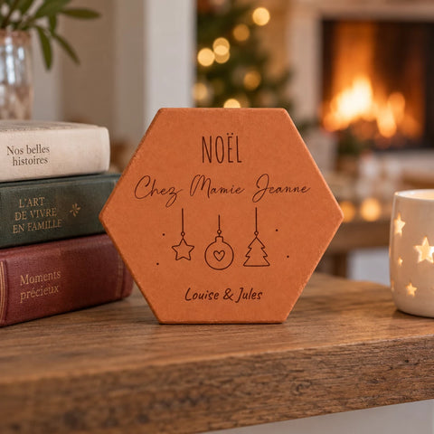 Tomette personnalisée « Noël chez Mamie Jeanne », Louise et Jules et décorations gravés, sur une table près de livres, d’une tasse et d’une cheminée allumée.