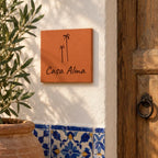 Plaque de maison personnalisée « Casa Alma » avec deux palmiers gravés, fixée près d’une porte en bois et de feuillage, au-dessus de carreaux bleus.