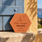 Tomette souvenir personnalisée « Côte d’Azur mon amour », Antibes, voilier et coordonnées gravés, adossée à un volet bleu, avec la mer au loin.