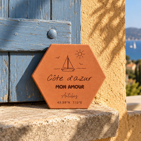 Tomette souvenir personnalisée « Côte d’Azur mon amour », Antibes, voilier et coordonnées gravés, adossée à un volet bleu, avec la mer au loin.