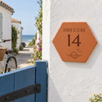 Plaque de maison personnalisée 14 chemin de la Mer, soleil et vagues gravés, fixée sur un mur blanc près d’un portillon bleu, avec la mer au loin.