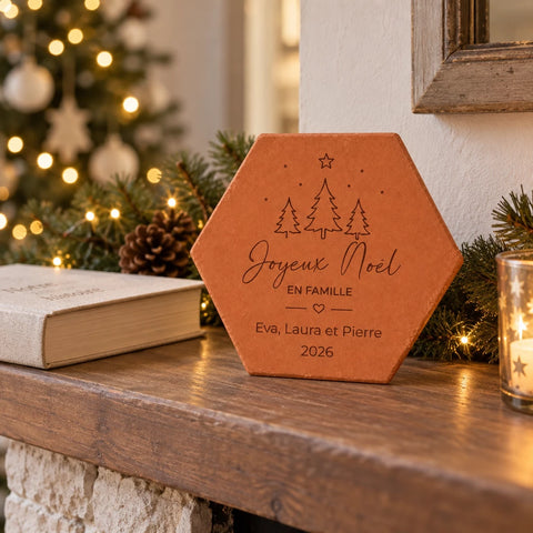 Tomette de Noël personnalisée « Joyeux Noël en famille », trois prénoms et sapins gravés, sur un manteau de cheminée près d’un livre et de branches illuminées.