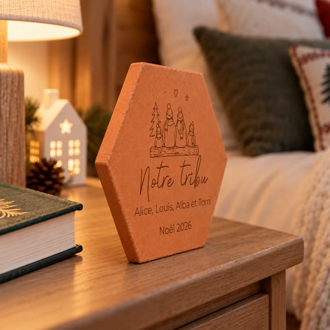 Tomette de Noël personnalisée « Notre tribu », quatre personnages et prénoms gravés, vue de côté sur une table de salon près d’un livre et d’une lampe.