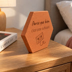 Terracotta tile engraved with “Parce que bien c’est pas suffisant”, an audio cassette and heart-shaped tape, seen from the side on the bedside table beside a book.