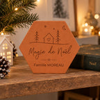Tomette de Noël personnalisée « Magie de Noël », Famille Moreau et petite maison gravées, près d’une maison lumineuse, de pommes de pin et d’un livre de Noël.