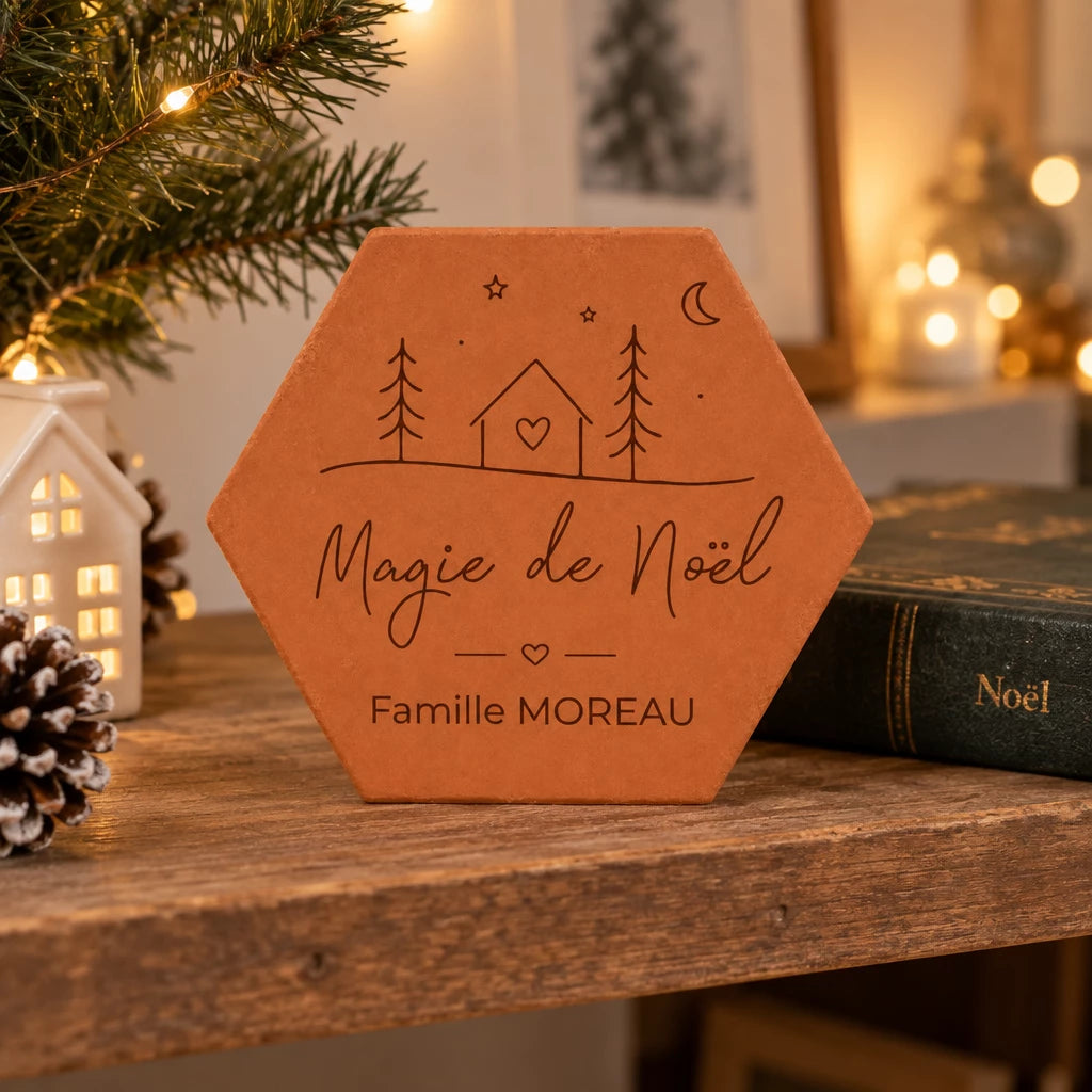 Tomette de Noël personnalisée « Magie de Noël », Famille Moreau et petite maison gravées, près d’une maison lumineuse, de pommes de pin et d’un livre de Noël.