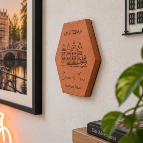 Tomette souvenir personnalisée Amsterdam, maisons, vélo, Emma et Tom et année gravés, vue de côté au mur près d’une plante et d’un album de voyage.