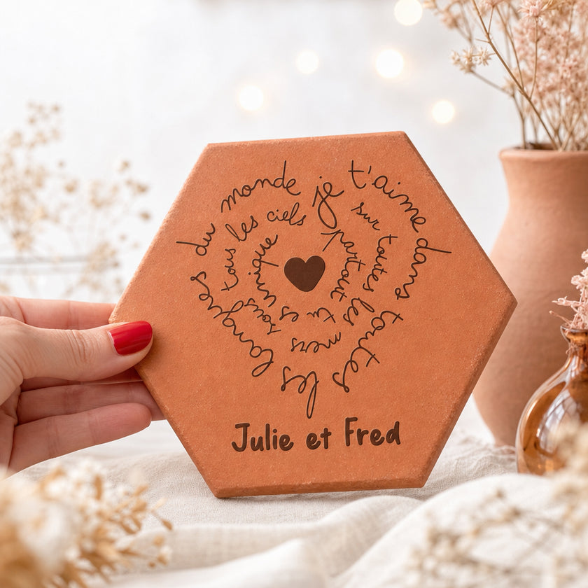 Personalised terracotta tile for the couple Julie and Fred, with words of love spiralling around a heart, held in a hand beside dried flowers and a vase.