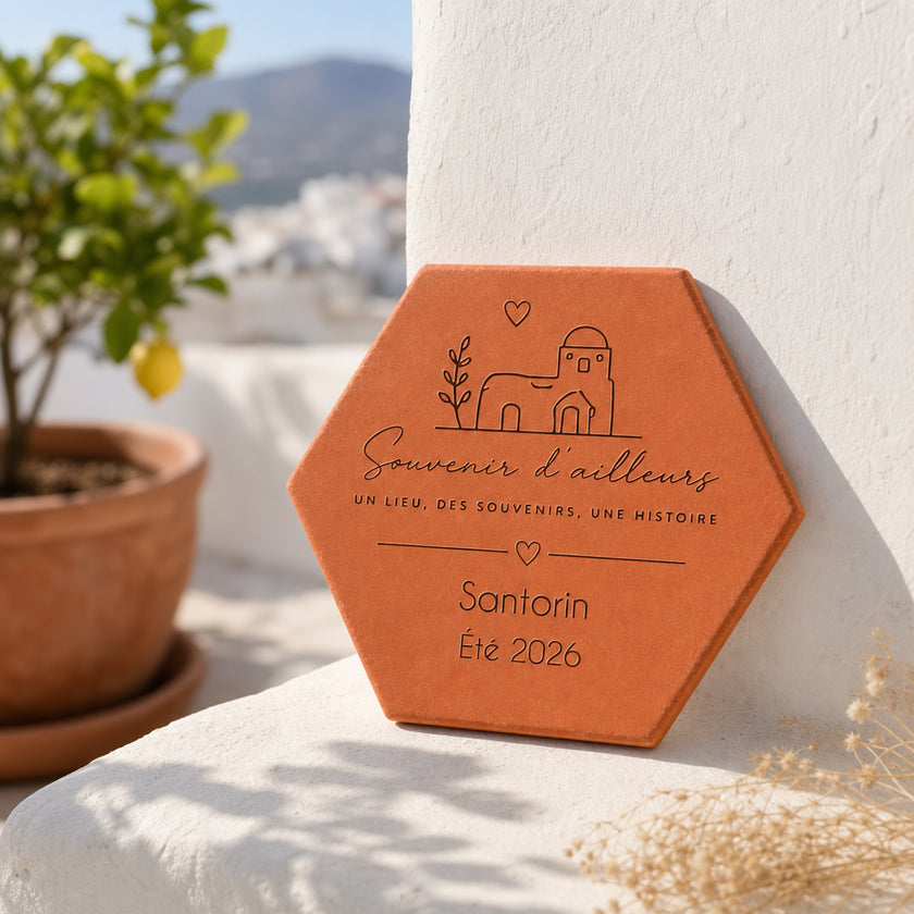 Personalised “Souvenir d’ailleurs” keepsake terracotta tile, engraved with “Santorin” and “été 2026”, leaning against a white wall beside a potted lemon tree.