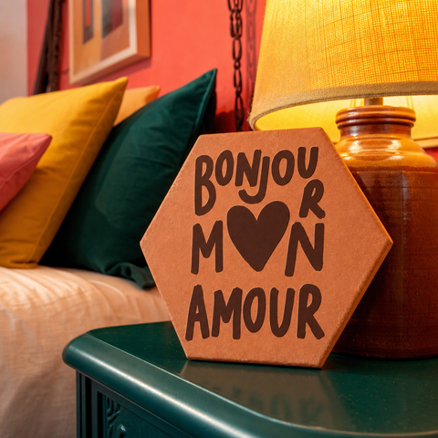 Tomette gravée « Bonjour mon amour » en grandes lettres avec cœur, dressée sur une table verte près d’une lampe et de coussins colorés.