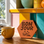 Tomette gravée « Bonjour », tasse à carreaux, croissant et cœurs, près d’une tasse jaune devant une crédence colorée.
