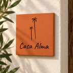Plaque de maison personnalisée « Casa Alma » avec deux palmiers gravés, vue de près sur le mur blanc à côté de feuillage.