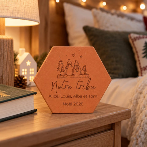 Tomette de Noël personnalisée « Notre tribu », quatre personnages et prénoms gravés, sur une table près d’un livre, d’une lampe et d’une petite maison lumineuse.