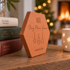 Tomette personnalisée « Noël chez Mamie Jeanne », Louise et Jules et décorations gravés, vue de côté près d’une pile de livres devant le sapin et la cheminée.