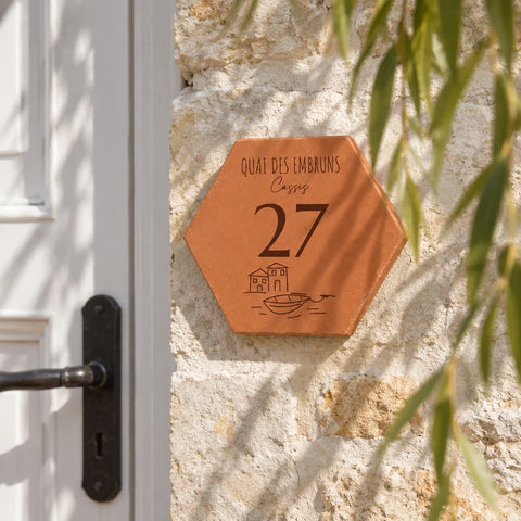 Personalised “Quai des Embruns, Cassis” house plaque, engraved with the number 27 and a small boat, fixed to a stone wall beside a white door.
