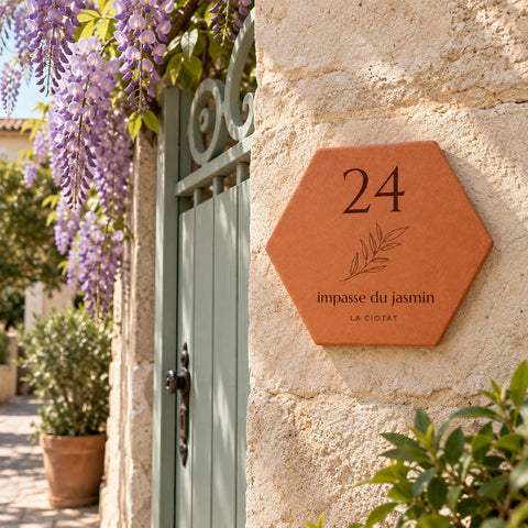 Plaque de maison personnalisée 24 impasse du Jasmin, La Ciotat et brindille gravés, fixée sur un mur en pierre près d’un portail et de fleurs mauves.