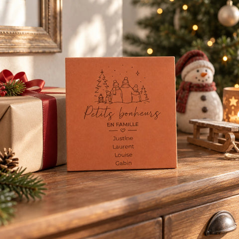 Carreau personnalisé « Petits bonheurs en famille », quatre personnages et prénoms gravés, sur un buffet près d’un cadeau et d’un bonhomme de neige décoratif.